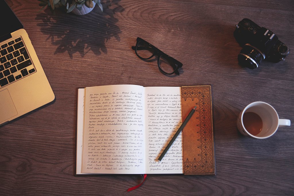 Vintage toned image of a open diary on a wooden desk, besides the laptop computer and a film camera. Coffee break for a couple of new lines.